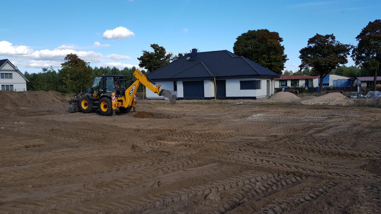 Żółta koparko-ładowarka JCB wykonuje prace ziemne na placu budowy nowego domu jednorodzinnego z ciemną dachówką, widoczne ślady opon na ubitej ziemi.