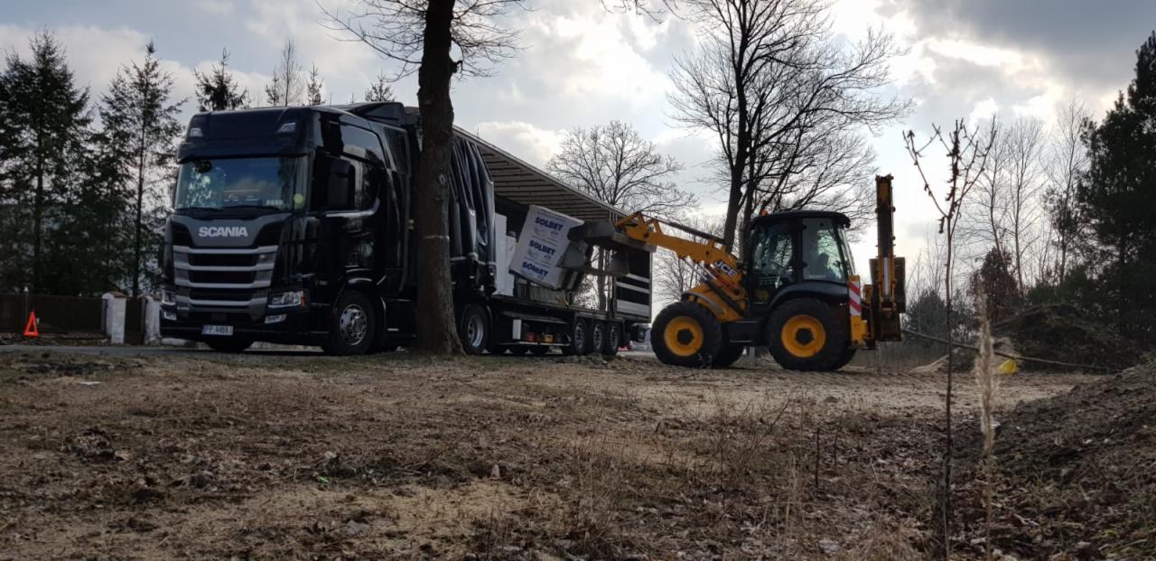 Żółta koparko-ładowarka JCB ładuje palety z kostką brukową Solbet na czarną ciężarówkę Scania na placu budowy w pochmurny dzień.