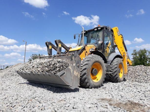 Żółto-czarna koparko-ładowarka JCB z pełną łyżką gruzu na tle błękitnego nieba z chmurami, stojąca na pryzmie gruzu.