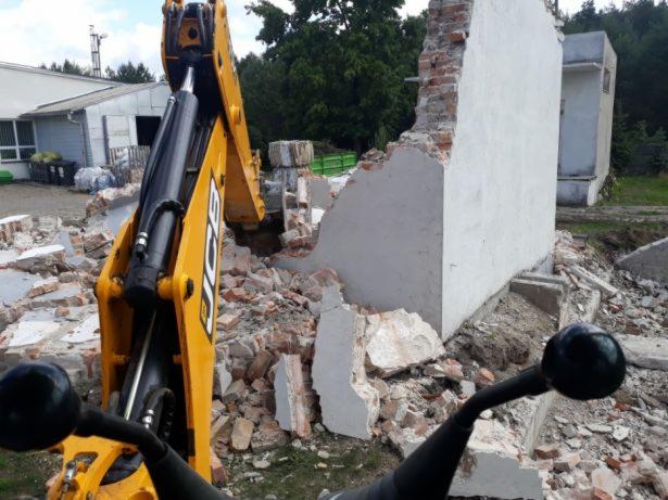 Żółta koparka JCB częściowo widoczna, gruz i fragment zburzonej, częściowo otynkowanej ściany budynku w tle
