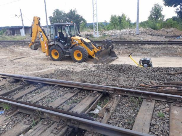 Żółta koparko-ładowarka JCB na torach kolejowych podczas prac ziemnych, ubijarka wibracyjna obok pryzmy tłucznia.