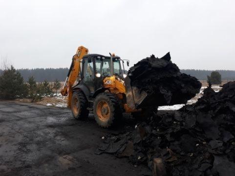 Żółta koparko-ładowarka JCB z pełną łyżką czarnej ziemi na tle pochmurnego nieba i lasu.