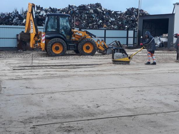 Żółta koparko-ładowarka JCB i operator zagęszczarki płytowej ubijający piasek na placu budowy, w tle hałda złomu i budynek gospodarczy.