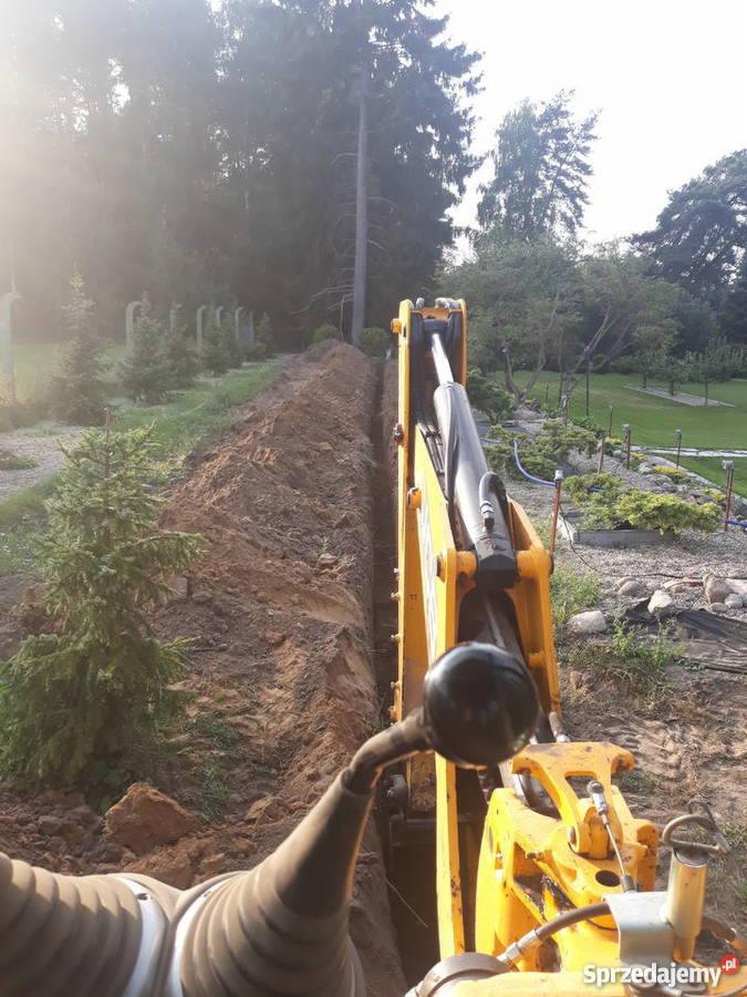 Żółta koparka w trakcie kopania wąskiego rowu w ziemi na tle zielonego ogrodu z drzewami i krzewami.