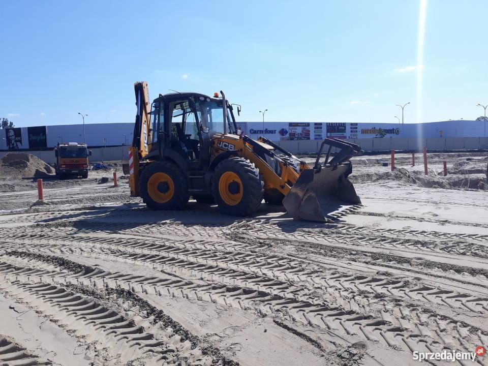Żółta koparko-ładowarka JCB na piaszczystym terenie budowy z widocznymi śladami opon, w tle budynek z szyldami sklepów i zaparkowana ciężarówka.