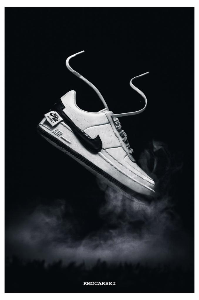 Czarno-białe zdjęcie lewitującego białego buta Nike Air Force 1 z czarnym logo na tle dymu i ciemnego tła.