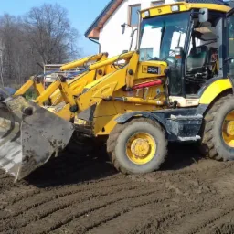 Żółta koparko-ładowarka JCB 3CX na placu budowy, widoczne świeże ślady opon na ziemi, w tle dom jednorodzinny w trakcie budowy.