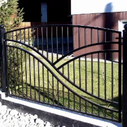 Czarny metalowy płot z dekoracyjnymi łukami i kulami na słupkach, na niskiej betonowej podmurówce z białymi kamieniami, na tle trawnika i budynku.