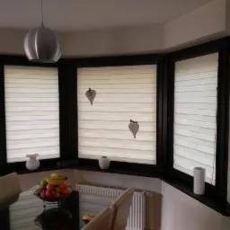 Trzy okna PCV z roletami plisowanymi w kolorze kremowym, dekorowane zawieszkami w kształcie liści, widok z jadalni z owocami na stole.