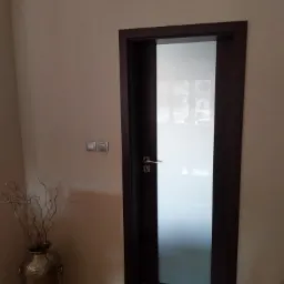 Nowoczesne drzwi wewnętrzne z matowym szkłem i ciemną ramą, widoczne z boku, z dekoracyjnym wazonem w tle