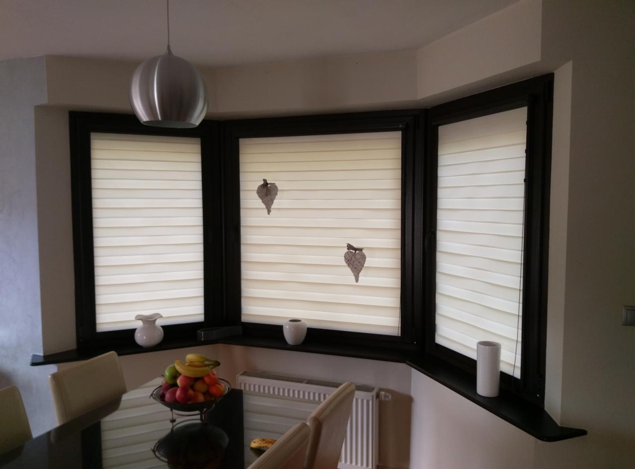 Trzy okna PCV z roletami plisowanymi w kolorze kremowym, dekorowane zawieszkami w kształcie liści, widok z jadalni z owocami na stole.