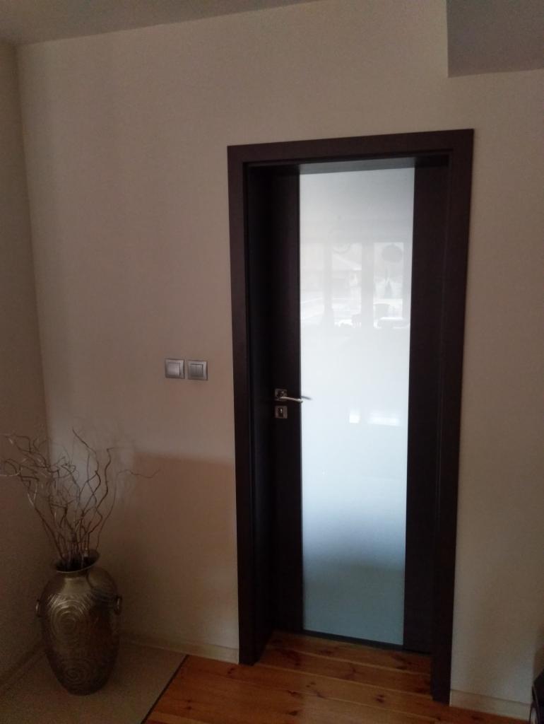 Nowoczesne drzwi wewnętrzne z matowym szkłem i ciemną ramą, widoczne z boku, z dekoracyjnym wazonem w tle