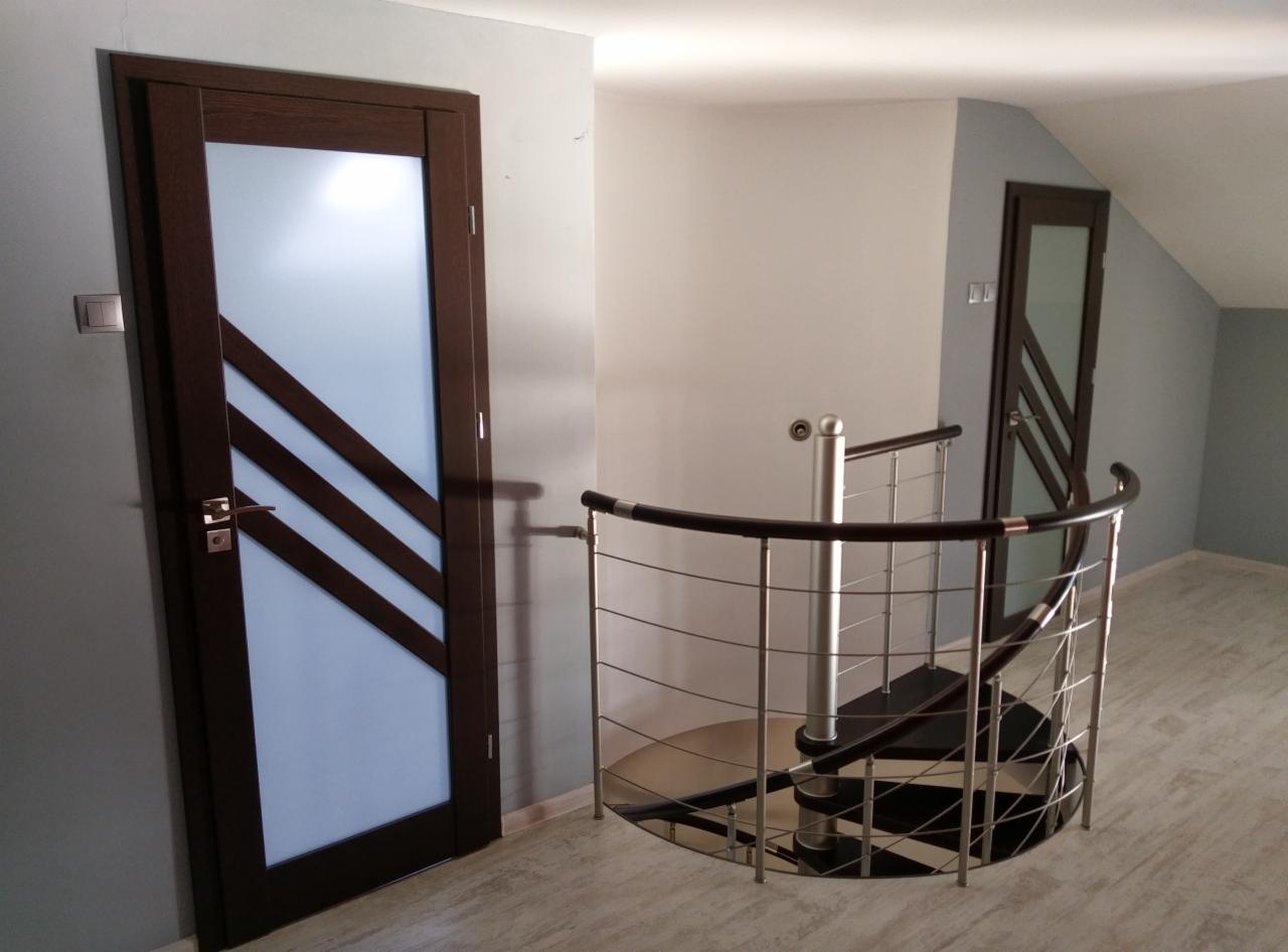 Wnętrze z drewnianymi drzwiami z matowym szkłem i trzema ukośnymi paskami, widoczne z poziomu schodów spiralnych z metalową balustradą w Radomsku.