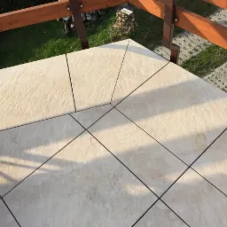 Jasne, kwadratowe płytki ułożone na tarasie pod kątem 45 stopni, z drewnianą balustradą w tle i widocznym fragmentem trawnika.