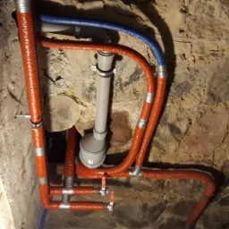 Instalacja wodno-kanalizacyjna z rurami w izolacji termicznej w piwnicy o kamiennych ścianach, widoczne zawory i kształtki.