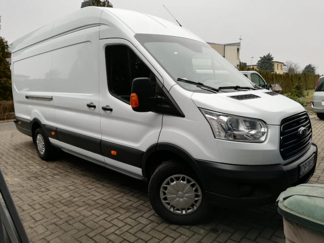 Biały, wysoki bus Ford Transit na brukowanym podjeździe, widok z boku, z widocznym numerem rejestracyjnym na przednim zderzaku.