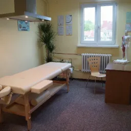 Jasny gabinet z beżowym stołem do masażu, biurkiem z modelem anatomicznym i dyplomami na ścianie, oświetlony lampą sufitową.