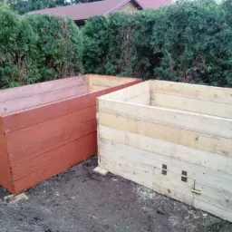 Dwa drewniane kompostowniki: jeden pomalowany na brązowo, drugi w naturalnym odcieniu drewna, ustawione na ziemi przed żywopłotem.