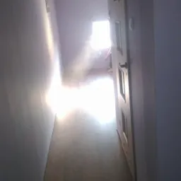 Korytarz z panelami podłogowymi w odcieniach brązu, ściany pomalowane na szaro-fioletowo, drzwi wewnętrzne z przeszkleniami, w głębi widoczne światło dzienne.