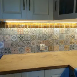 Wzorzyste płytki ceramiczne nad drewnianym blatem kuchennym, oświetlone listwą LED, z widocznym gniazdkiem elektrycznym i fragmentem białych szafek.