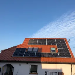 Dom jednorodzinny z panelami słonecznymi na dachu z czerwonej dachówki, widoczne okna dachowe i błękitne niebo z chmurami.