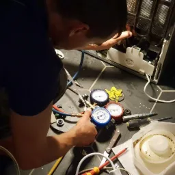 Serwisant w trakcie naprawy lodówki, podłącza manometry do układu chłodniczego, widoczne narzędzia i elementy demontowane z lodówki na podłodze z ciemnych płytek.