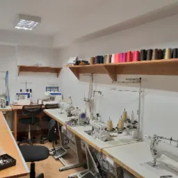 Wnętrze szwalni z kilkoma maszynami do szycia JUKI na białych stołach, półki z kolorowymi nićmi, miarka krawiecka i pudełko z guzikami na stole.