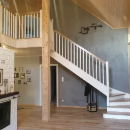 Wnętrze domu szkieletowego z drewnianymi schodami z białymi balustradami, widok na antresolę i kominek wykończony białymi kaflami, ściana pomalowana na szaro, oświetlenie w stylu industrialnym.