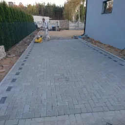 Układanie szarej kostki brukowej w jodełkę na podjeździe, z ciemnymi akcentami wzdłuż krawędzi, pracownik zagęszcza powierzchnię mechaniczną ubijarką.