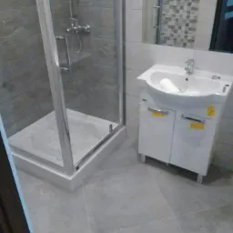 Wykończona łazienka z kabiną prysznicową z chromowanymi okuciami, białą szafką pod umywalką z ręcznikiem i szarą glazurą na ścianach i podłodze.