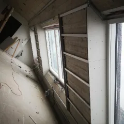 Poddasze w trakcie izolacji, widoczne okna, płyty izolacyjne z metalowym stelażem, surowy betonowy strop i przewody elektryczne.