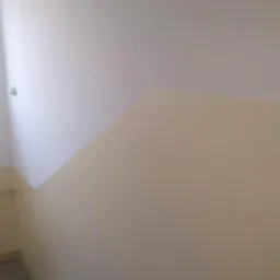 Klatka schodowa po malowaniu, z szarymi schodami i dwukolorowymi ścianami: górna część biała, dolna kremowa, widoczny fragment poręczy.