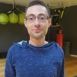 Mężczyzna w okularach i bluzie z kapturem w tle sali z workiem bokserskim, piłkami i sprzętem do ćwiczeń.