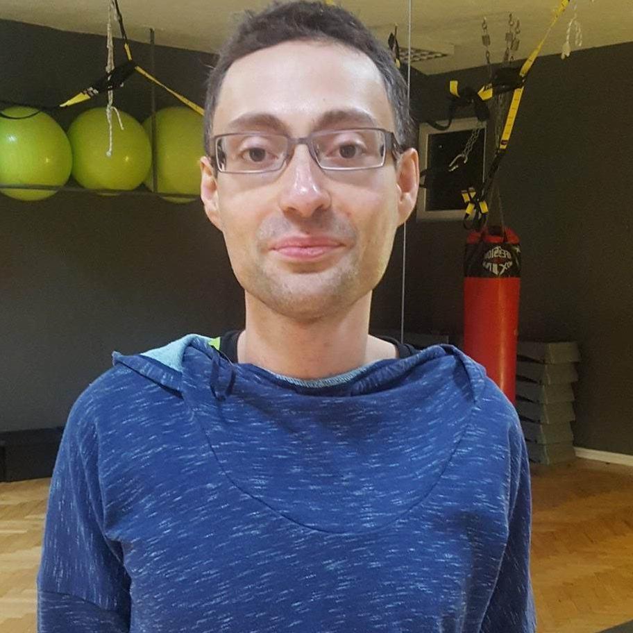 Mężczyzna w okularach i bluzie z kapturem w tle sali z workiem bokserskim, piłkami i sprzętem do ćwiczeń.