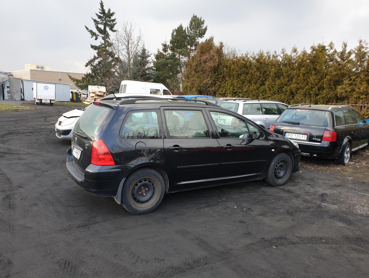 Czarny samochód kombi Peugeot 307 na placu z innymi pojazdami, widoczne ślady rdzy na felgach i błotniste podłoże.