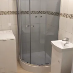 Ukończona łazienka z kabiną prysznicową narożną, umywalką na szafce i pralką, wykończona białymi płytkami z dekoracyjnym pasem mozaiki.