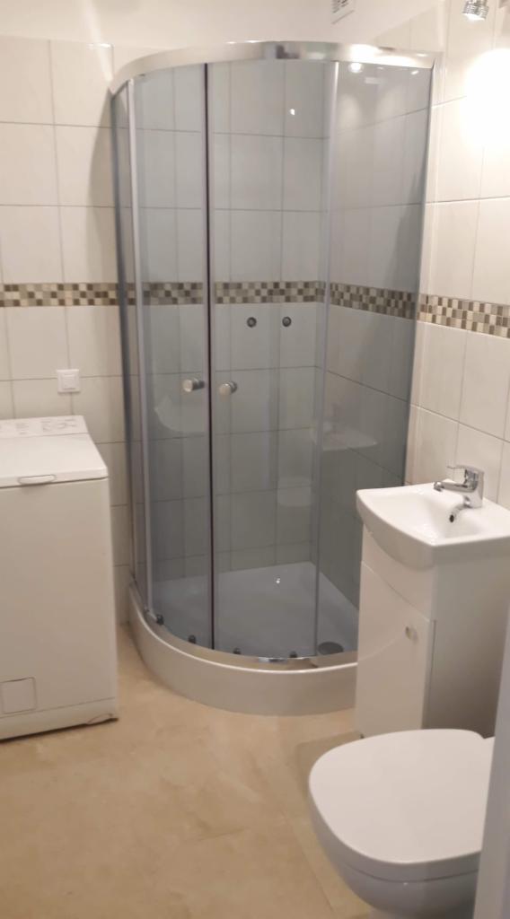 Ukończona łazienka z kabiną prysznicową narożną, umywalką na szafce i pralką, wykończona białymi płytkami z dekoracyjnym pasem mozaiki.