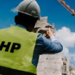 Mężczyzna w białym kasku i jaskrawożółtej kamizelce z napisem BHP, stojący tyłem na tle budowy i błękitnego nieba z chmurami. Widoczny żuraw budowlany.