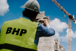 Mężczyzna w białym kasku i jaskrawożółtej kamizelce z napisem BHP, stojący tyłem na tle budowy i błękitnego nieba z chmurami. Widoczny żuraw budowlany.