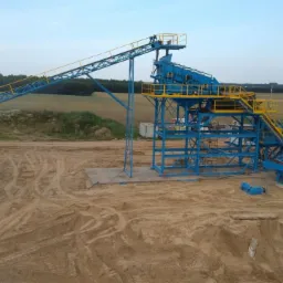 Kompleksowa instalacja do sortowania kruszywa w kolorach niebieskim i żółtym, z widocznym podajnikiem taśmowym i konstrukcją wsporczą, na tle pola i lasu.