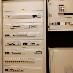 Rozdzielnica elektryczna z wieloma zabezpieczeniami nadprądowymi i różnicowoprądowymi, widoczne przewody w górnej części, obok otwarta szafka elektryczna.