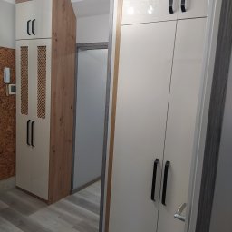 REFIT ADAM KORODY - Biała szafa z czarnymi uchwytami i ażurowym frontem w przedpokoju, widoczny fragment korkowej ściany i drzwi do innego pomieszczenia.