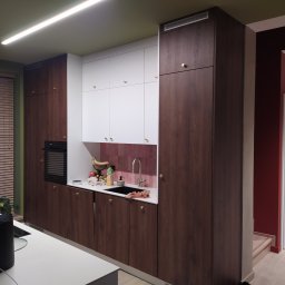 REFIT ADAM KORODY - Nowoczesna kuchnia z drewnianymi szafkami dolnymi i białymi górnymi, złotymi gałkami, różowymi płytkami nad blatem i owocami w zlewie.