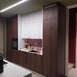 Nowoczesna kuchnia z drewnianymi szafkami dolnymi i białymi górnymi, złotymi gałkami, różowymi płytkami nad blatem i owocami w zlewie.