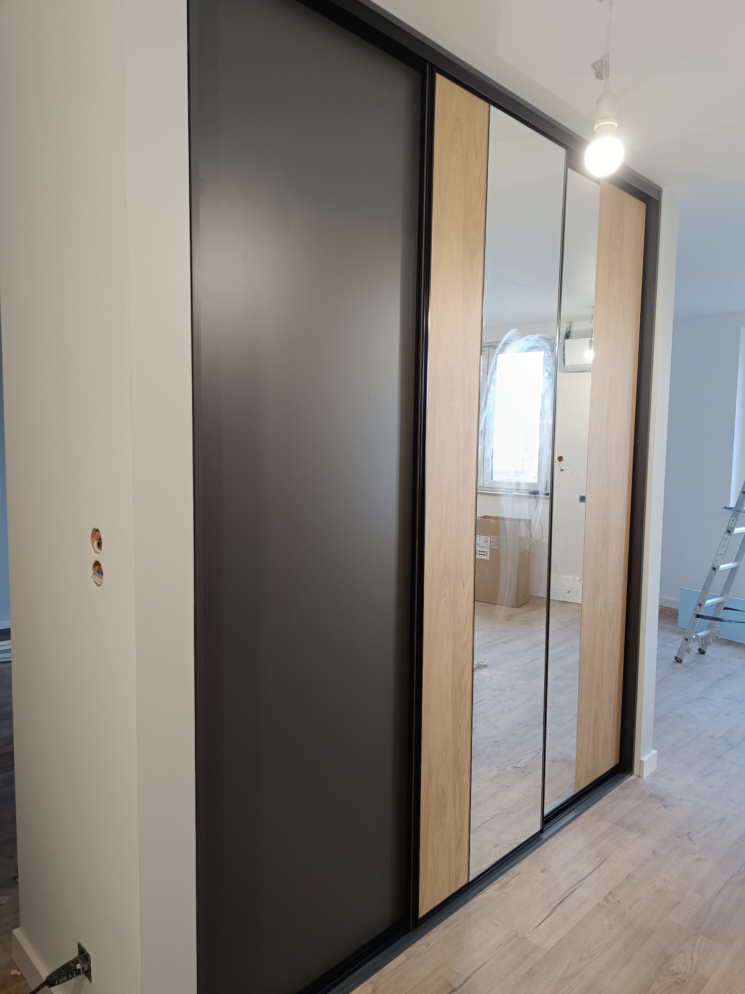 Nowoczesna szafa wnękowa z przesuwnymi drzwiami: czarny panel, lustro i drewnopodobny akcent, widoczne przygotowania do montażu, drabina w tle.