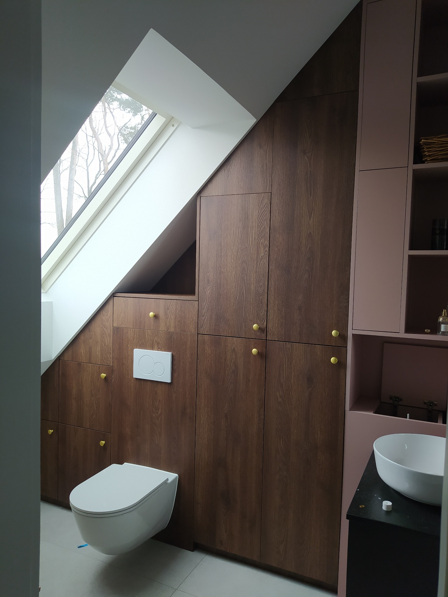 Łazienka na poddaszu z zabudową z ciemnego drewna pod skosem, dopasowaną do okna dachowego, z toaletą podwieszaną i różowymi szafkami z umywalką.