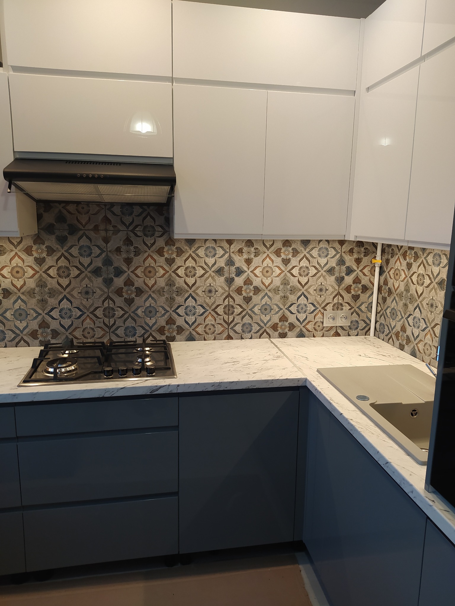 Nowoczesna kuchnia z białymi szafkami górnymi, szarymi dolnymi, marmurowym blatem i płytkami w stylu retro nad kuchenką gazową.