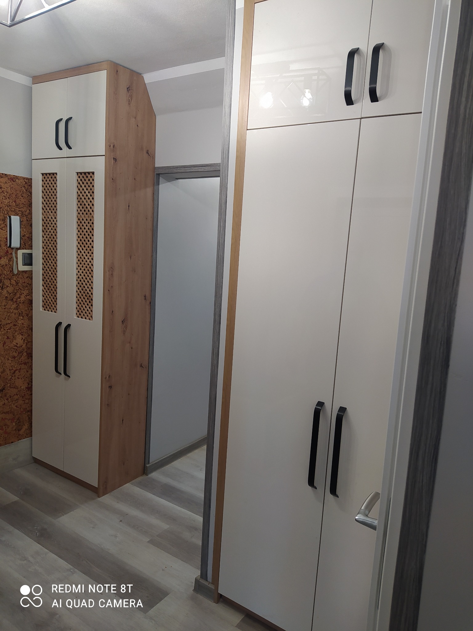 Biała szafa z czarnymi uchwytami i ażurowym frontem w przedpokoju, widoczny fragment korkowej ściany i drzwi do innego pomieszczenia.