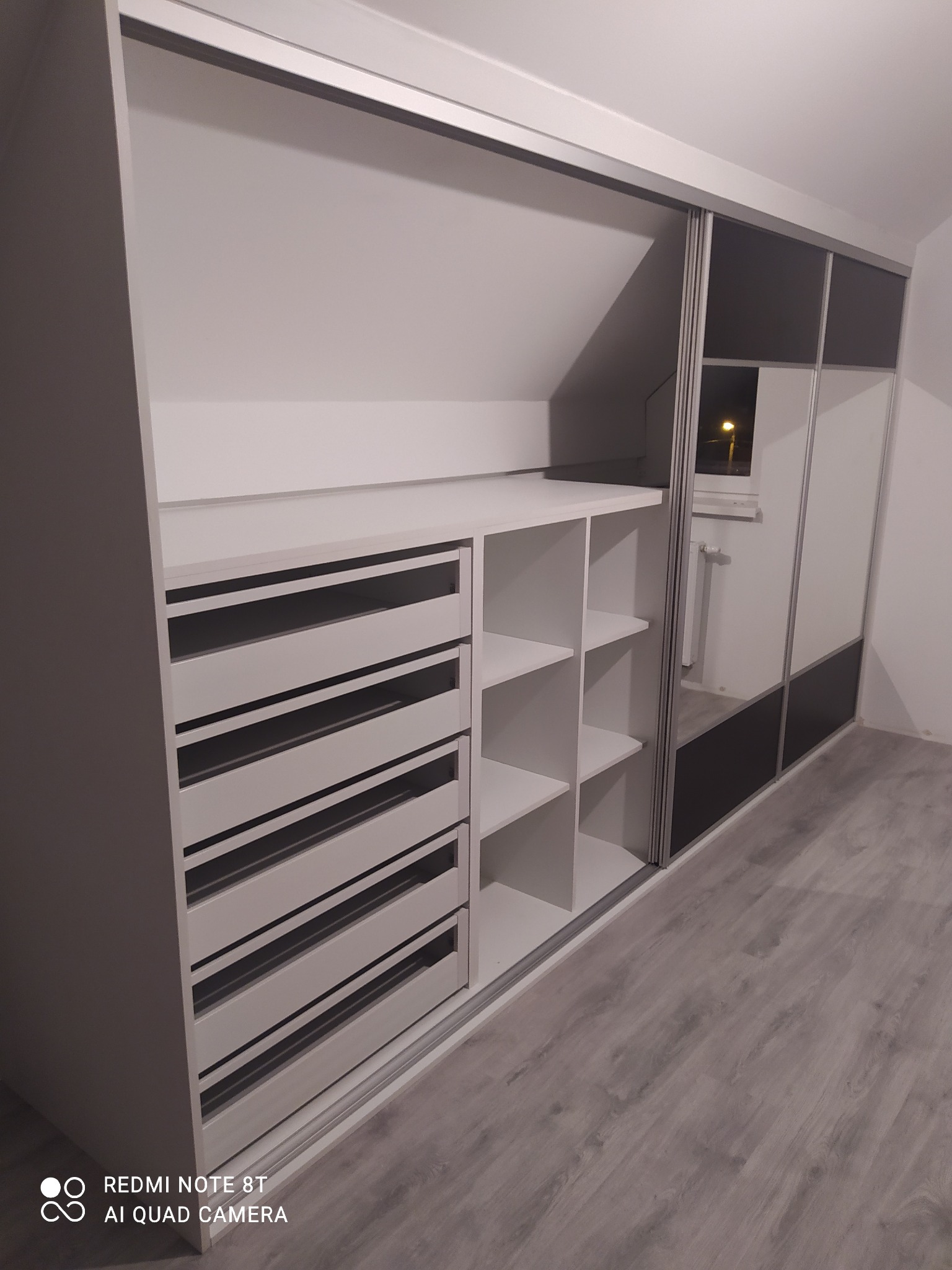 Biała szafa wnękowa z przesuwnymi drzwiami z lustrami i czarnymi akcentami, widoczne szuflady i półki, zamontowana na poddaszu.