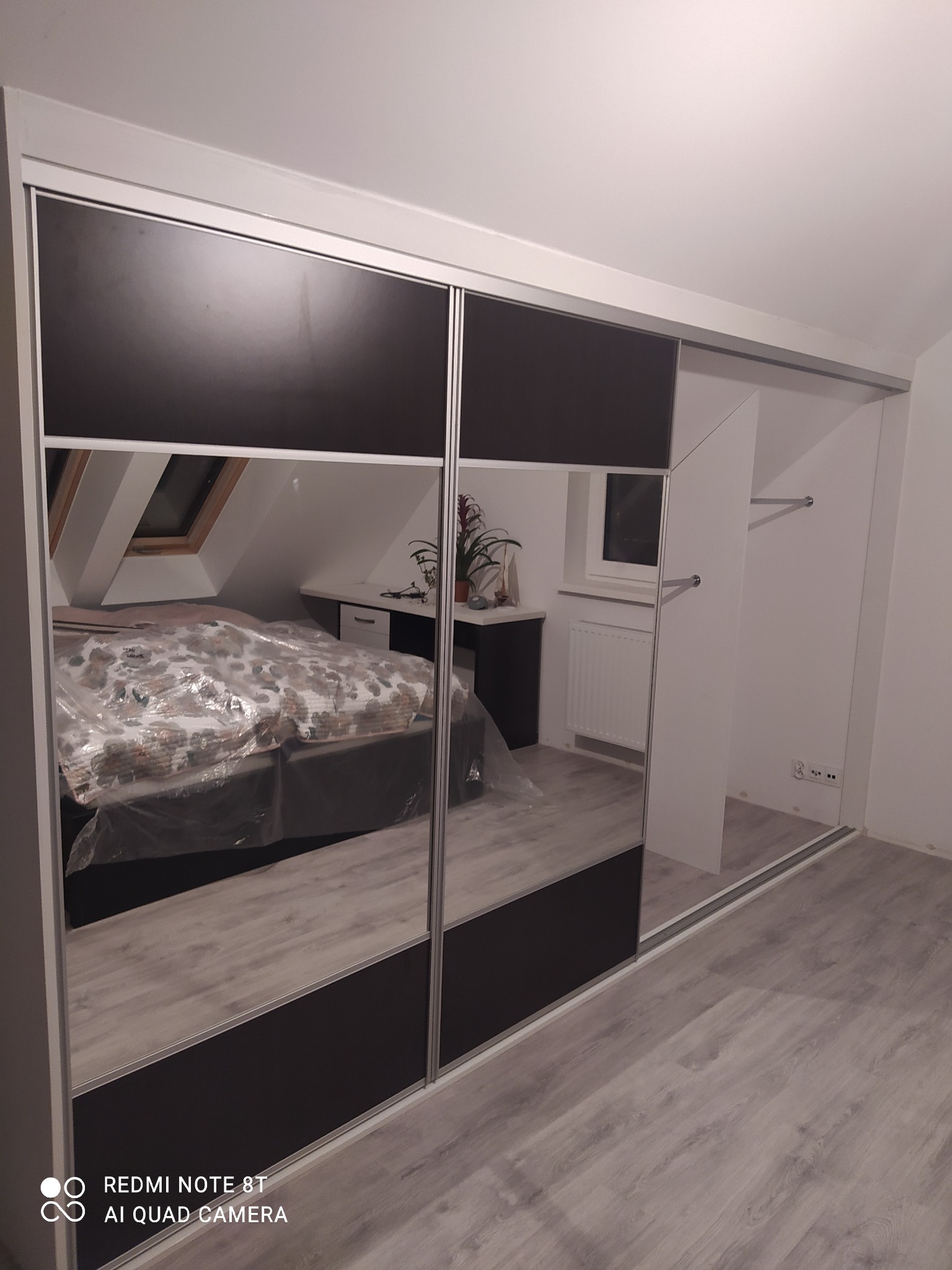 Czarno-biała szafa przesuwna z lustrzanymi drzwiami, odbijająca pokój na poddaszu z oknem dachowym i łóżkiem przykrytym folią ochronną.